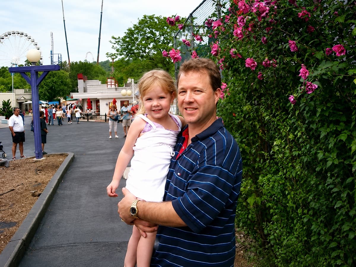 Peter Munro Roberts in a navy striped polo holding a young girl at an amusement park, with a Ferris wheel and rose bushes in the background.