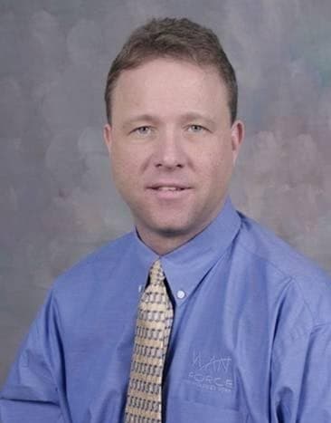 Studio portrait of Peter Munro Roberts in a blue shirt bearing the Wanforce logo, with a patterned tie, against a gray backdrop.